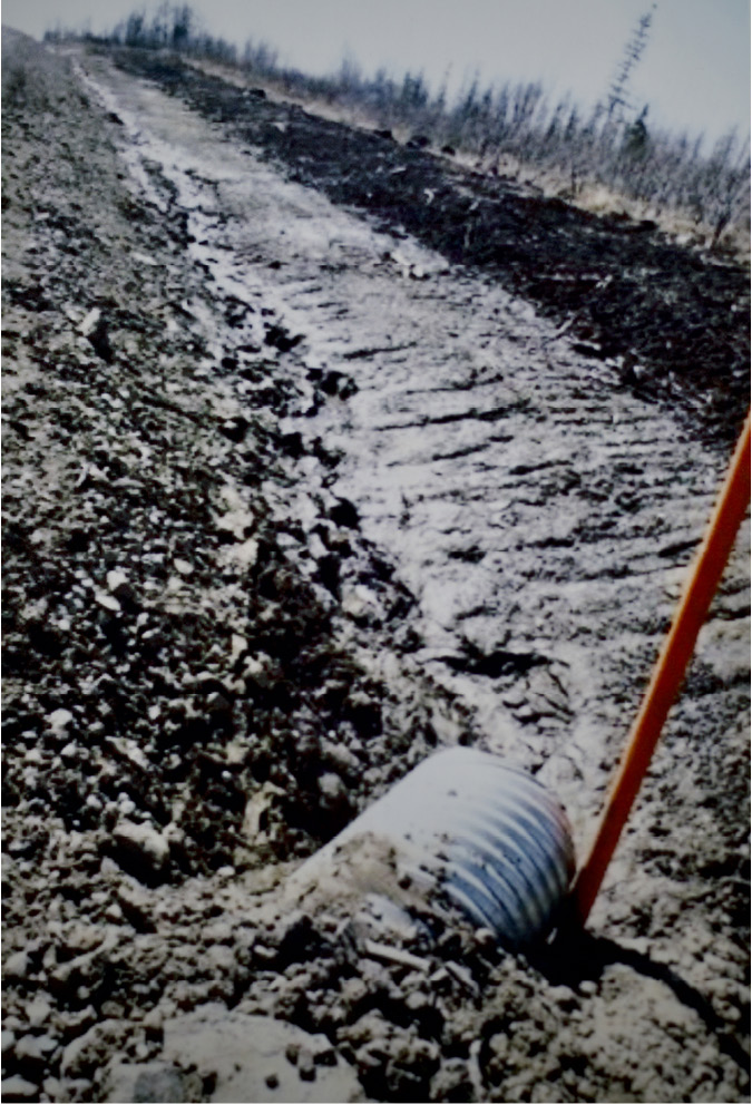 In the foreground is a large industrial steel pipe vertically shooting out into the ground of the Brassy Creek shores near hydroelectric production areas. There is a vertical flow meter, illustrated by a long thin tube installed next to the pipe. This measure the quality of the water flow that is moving from the side to create a dugout with low bank as the water moves.