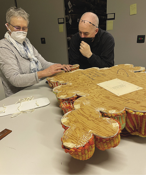 Photo: Two people wearing masks look at the wooden backing of something on a high table.
