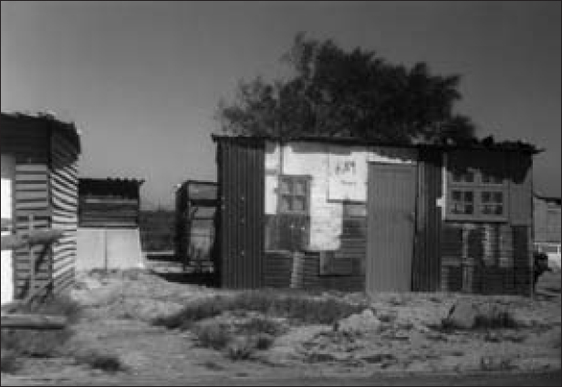 A view of a small house made of corrugated iron, with a door, two windows and a flat roof, located on dry land. Three similar structures are seen around the house.