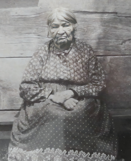 Black and white historical photograph. An elderly Indigenous woman is seated in front of a wooden wall. She is wearing a long dark dress with dots and a decorated fringe, and a long-sleeved shirt with a floral pattern. Her grey hair is pulled back from her face.