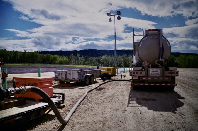 Dawson Creek to the left of the image, soft rolling and tree covered hills provide the background. In the foreground is a larger tanker truck with a hose joined to other equipment.