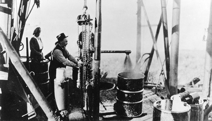 This photo shows Charles Taylor turning a valve to release steam and oil into a large barrel at the newly built Norman Wells refinery. A woman stands behind him. Taylor is in shirt-sleeves and a vest with a hat. He was one of the partners in the Northwest Company in 1919-21, and died under mysterious circumstances several years later.