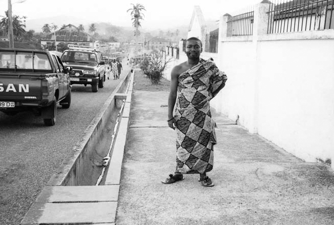 A black and white photograph of Nana Fredua Agyeman standing in a African dress. On the right side, there are some cars parked and a drain is running through the road.