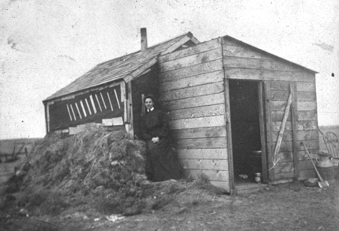 A black and white photo of a wood plank shack with a woman and haystack. The shack has an entryway or mud room with a roof that slants down at an angle from left to right. The door is open. A shovel leans against the wall to the right of the door. To its right there is a large container, perhaps a water container or a churn. The entrance opens into a frame shack with a peaked roof. The side appears darker than the planks. Perhaps it was covered with tarpaper. There is a stove pipe protruding from the roof. There is a small haystack piled about halfway up the side wall. The one-room shack is a bit wider than the entryway, and a woman stands in the corner formed by the shack and the protruding entryway, next to the haystack. She wears a dark long dress with long sleeves and a white collar. Behind the shack we can see the flat prairie horizon. 