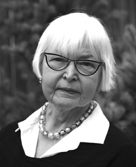 Black and white photograph: A portrait of an older woman with white hair, wearing stylish glasses, pearls and a white blouse. She is looking directly at the camera.