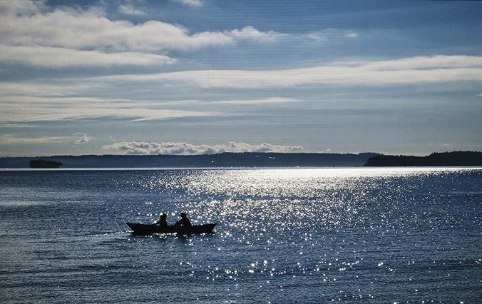 A large body of water is in the foreground, with light on the water both casting shadows and sparkling. Two people are seated in a boat, one is rowing, shaded in the view with just low rolling hills and a few wispy clouds in the background.