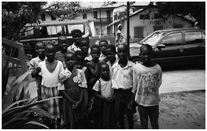 A black and white photograph of a group of Manya Krobo schoolchildren, standing next to a road with four wheelers parked. Houses are seen on the other side of the road.