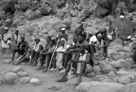 Elderly men seated on an arrangement of stones. Some of them hold sticks.