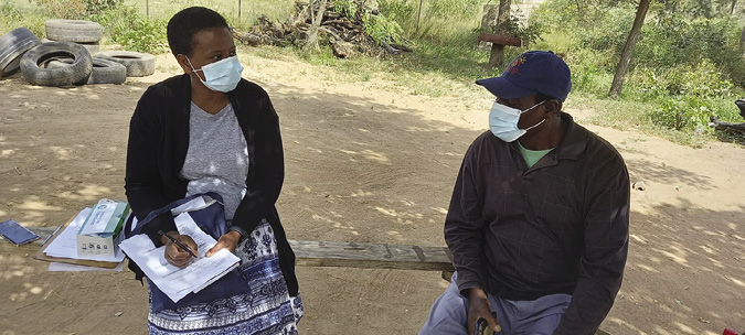 Colour photograph: Photo of a woman researcher with pen and clipboard and a male farmer, both wearing Covid masks, sitting on a bench outdoors, talking. In the background are trees, brush, a pile of old truck tires.
