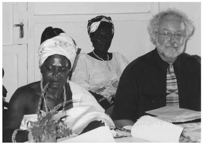 A black and white photograph of Doctor Don Ray, with an elderly woman Manye Nartekie, Deputy Paramount Queenmother, sitting at a desk with files and documents. Another woman is sitting behind them. Both African women are wearing traditional clothing, one with a gele and the other with a head tie.