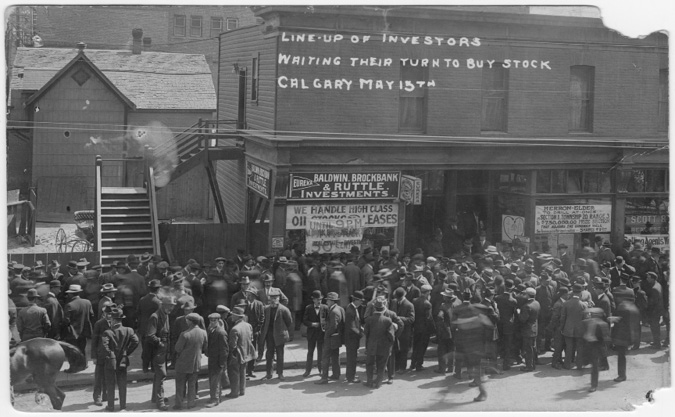 Figure 5-4 “Investors Waiting to Buy Oil Stocks in Calgary, Alberta.” Black and white photograph taken outside 801, 1st Street SW where a substantial crowd of people overwhelm the sidewalks and mill on in the street in front of two businesses that are selling stock in oil companies. In the front windows of two establishments are handmade signs advertising the stocks for sale. In the shop window of Baldwin, Brockbank and Ruttle Investments is a sign that reads “We Handle High Class Oil Stocks and Leases.” Another sign partially covers this first one, advertising a discount on some shares to fifty cents until 9 pm. In the window on the right of the door, a picture of the Herron-Elder’s “Heart of the Oil Fields” symbol s visible. Over the larger window is a sheet with a hand-painted sign for Herron-Elder stock.