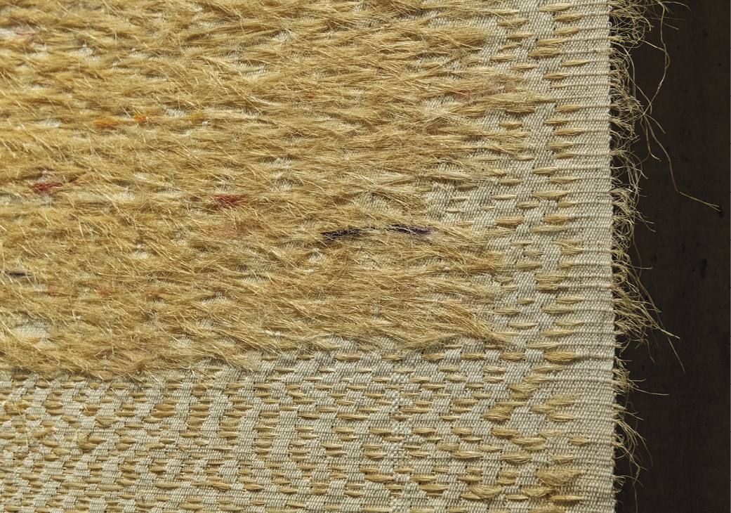 Photo: detail of a horizontal piece with straw-coloured threads going throw cream material. Chunks are messy and above the material, with a fringe coming off the right edge. 