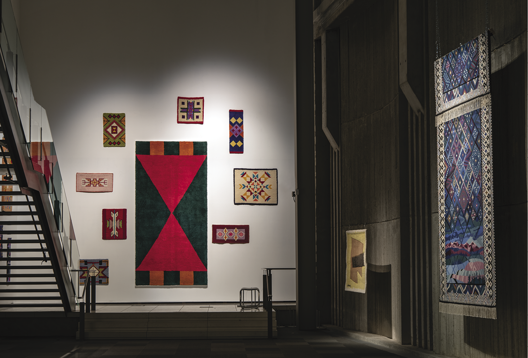 Photo: a very tall white wall with one large red-green hanging surrounded by eight much smaller hangings with complex patterns. A staircase leads up along the left. A grey-black wall on the right has two hangings — one tan coloured and tiny, the other extra large, and in shade of blue with a complex pattern. 