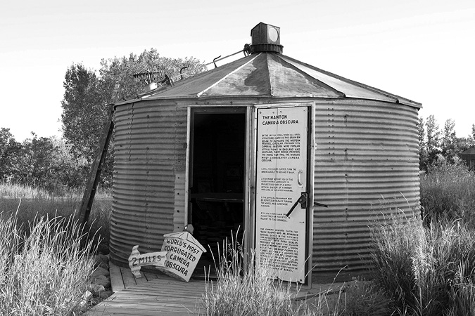 Black and white photograph, a corrugated iron tank with an open door, a conical roof, and a box with lens at the top of the roof; at the bottom of the door signs advertise the structure as “world’s most corrugated camera obscura”.