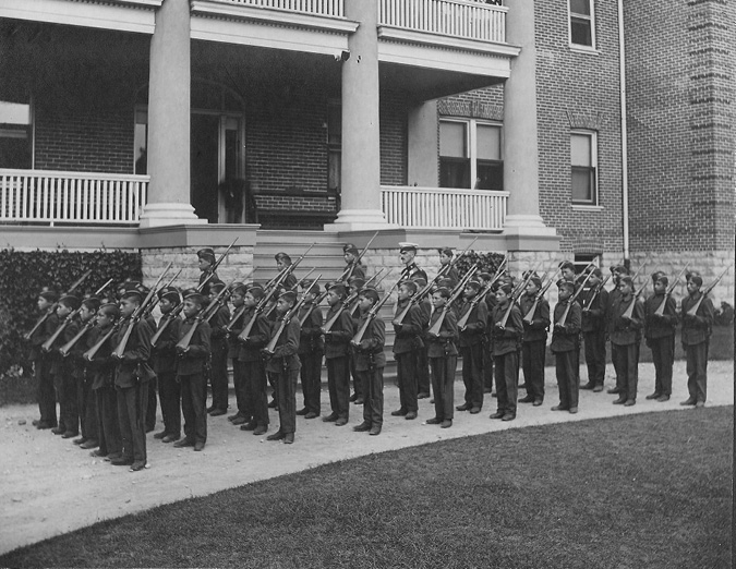 Black and white photograph: Nine rows of male Indigenous students in cadet uniforms posed on the driveway in front of the steps of the Mohawk Institute. There are approximately sixty students in total.  Their male teacher is also in the photograph.  The students are standing, each holding a firearm on their left shoulder.