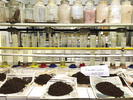 Colour photograph: Soil samples on pieces of newspaper on a shelf in a laboratory with scientific bottles and beakers behind the samples.