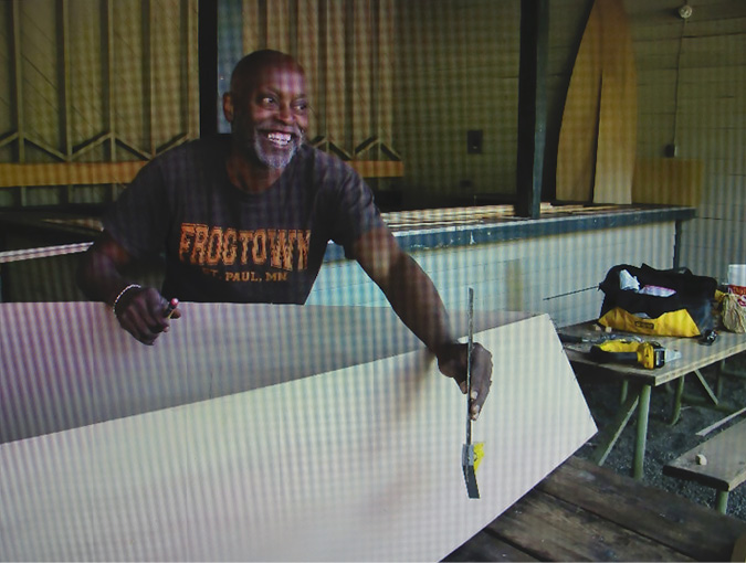 . Seitu Jones, facilitator of a creative advocacy art project, is smiling broadly as he stands behind a large white boat, holding a leveler/measuring tool. Other building supplied are place on top of a picnic table next to Jones, while other parts of the boat lean against a stage in the background.