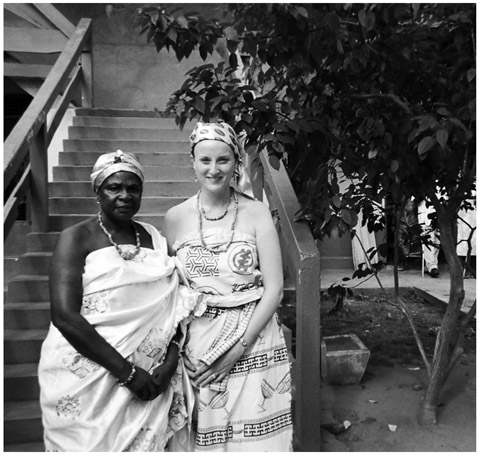 A black and white photograph of Manye Nartekie, Deputy Paramount Queenmother, with Sherri Brown, standing outside the Manya Krobo Queenmothers Association Resource Centre. Both women, African and American, are in traditional African attire.