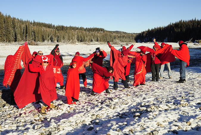 A group of people, wrapped in red fabric, are in the foreground standing on the banks of a river with small rocks. The group are holding their arms in the air, some are crouched down, one is holding a red umbrella. The low hills in the background frame the image.