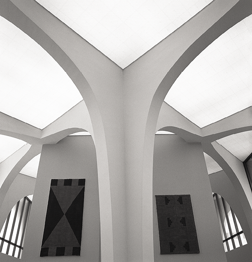 Black and white Photo: rounded wide open entry ways in front of white walls with rugs hanging on them between white panel lights.