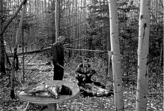 The image is a black and white photograph depicting a forest scene. In the foreground, there is a circular table displaying the body of a skinned bear, the head remains covered in fur. To the right, two men are visible in the woodland setting. One man (Earnie Lacorde) stands, holding a knife, while the other (Alex Boucher) crouches beside the remains of skinned bear, holding the bears head in his hand with a cigarette in his mouth. Both men smile gently, appearing amused. The forest is dense with tall, slender trees, most of which have smooth bark. The forest floor is covered with fallen leaves, adding texture to the scene. The overall mood is light and centered on the interaction between humans and nature.