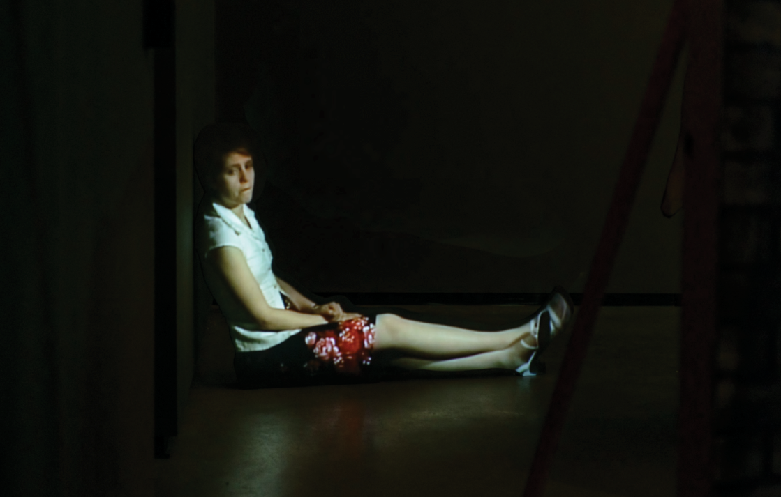 Colour photograph: A projection of a woman seated on the floor with her back against the wall, looking towards the viewer.
