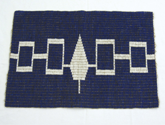 Colour photograph. Photo of a wampum belt, with the symbol of the Haudenosaunee Confederacy. The belt is mostly purple, with a white design running through the centre. This is a line, with four rectangles along it, two on either side. In the middle there is a representation of a pine tree, indicating the Great Tree of Peace.