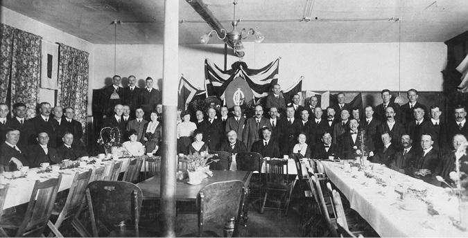 This is a photo of a Joint Industrial Council meeting around the time they were established by Imperial Oil (following the lead of Jersey Standard). Council members for the Sarnia refinery are gathered around several long tables with white tablecloths set for a formal dinner. Flags hang in the background. There are about five women in the group of several dozen managers and workers. All are dressed in business attire.