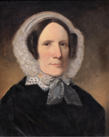 Colour painting. Head and shoulders portrait of a woman with black hair and brown eyes, wearing a black coat and a white head covering tied with a wide ribbon below her chin. Her hair is formed into a large curl on each side of her head. 
