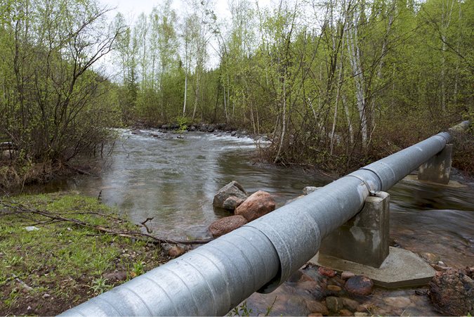 A large steel pipe crosses a rocky stream, propped up by cement structures. Lining the stream are trees, it is fall and the leaves have fallen from the, now bare, trees.