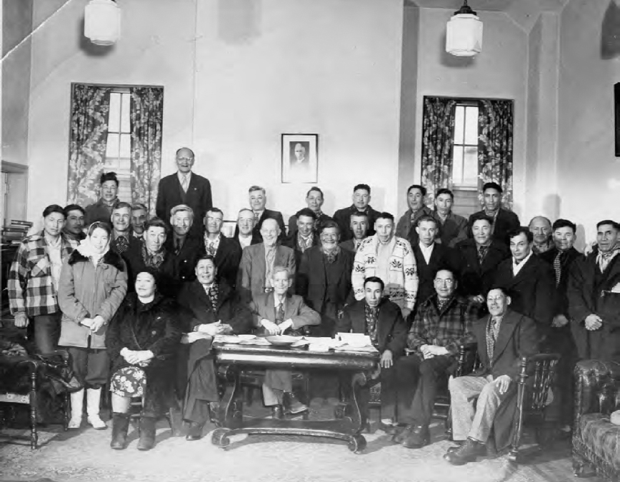 A photograph of a group of people in formal dress. Two windows, ceiling lights and a wall can be seen in the background.