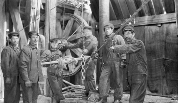 Figure 1-6 “Drillers at Dingman #1” Black and white picture of six oilfield workers in the drill shack of the Dingman #1 well, near the walking beam of the cable tool rig. All six workers are wearing coveralls, a cap and leather gloves. Driller Joseph Brown stands on the extreme right of the picture, with his left hand on the drilling cable running to the Bull Wheel.