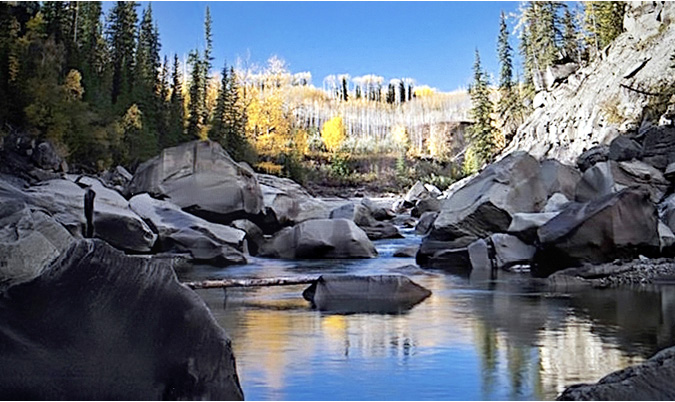 Large boulders and felled trees sit in a narrow bank of the Upper Kiskatinaw River, the steep slopes proving the perfect slide. In the background, evergreen and deciduous trees cover the soft mountains. A beautiful and ominous view, leaving the viewer wondering where the water will flow through this newly shaped landscape?