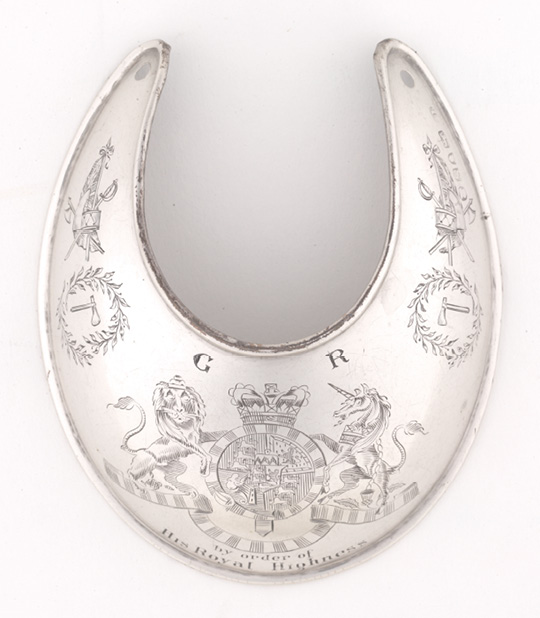 Colour photograph. A silver gorget on a white background. It is elaborately engraved. The British coat of arms is in the centre, with the letters G.R. above it. On either side there is a tomahawk surrounded by wreaths of leaves, and a composite image of a sword, battle axe, drum and British flag.