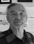 Black and white photograph: Portrait of a man wearing a collared shirt with some frames behind him on a wall.