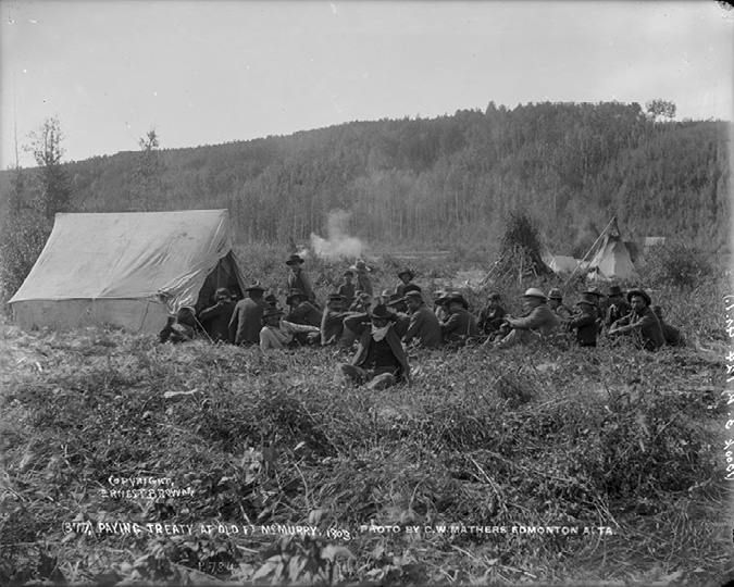 The photograph depicts an outdoor scene with several people gathered in a clearing surrounded by dense forest. A large canvas tent is prominently positioned on the left side, with a smaller tent visible in the background to the right. The people, mostly men, are sitting or crouching on the ground in a semicircle, wearing wide-brimmed hats and dark clothing. The vegetation around them appears wild and untamed, with a mixture of grass and shrubs. The distant background is dominated by a thick expanse of trees that stretches across the horizon. The overall atmosphere suggests a meeting or gathering, with attention focused towards a central figure not visible in the image.