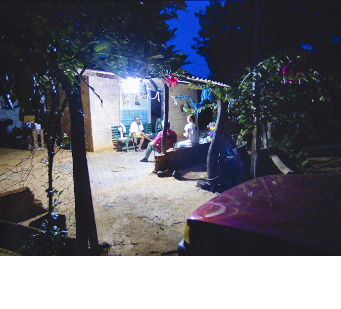An interview is occurring under a sheltered patio of a dwelling. The earth is cracked and dry on the ground at this dusk darkening sky, a window in the house behind the patio lights up the patio shelter. Under the shelter, in the light from the house, are benches facing one another.  The interviewers on one side and the interviewees on the other.