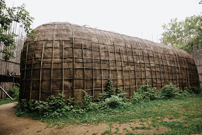 Colour photograph. Photo of a reconstructed longhouse. The longhouse is a large, long building with a domed roof. It is constructed from the poles of saplings and is covered in tree bark. A stockade of tall poles made of tree trunks can be seen behind it.
