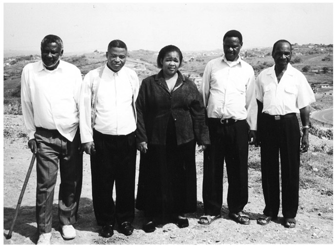 A black and white photograph of four men standing on a field with a woman in the centre. All the men are wearing white shirts and black trousers and the woman is wearing black top over black gown.