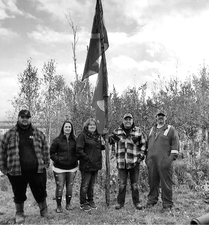 The black-and-white image shows five people standing outdoors on grass, with a backdrop of tall trees and a cloudy sky. From left to right, a man in a plaid jacket and a baseball cap, a woman with long hair in a puffer jacket, another woman in a similar jacket holding a tall flagpole with several flags, a man in a plaid jacket with a cap also holding the flagpole, and another man wearing industry coveralls with a baseball cap. The group is positioned in front of dense foliage, with the flags prominently visible above their heads.