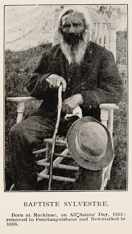 Black and white historical photograph. Portrait of an elderly man with a long beard sitting in a wooden chair. He is wearing a coat and holding a cane and handkerchief. There is a hat sitting on his knee. A caption for the photograph is at the bottom.