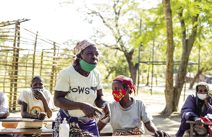 Colour photograph: Photo of a woman outside under a tree wearing a Covid mask; five other people listen to her speaking.