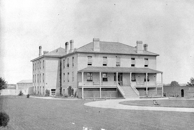 Black and white photograph: Two storey brick school building with a basement from the 1880s. This is the second set of Mohawk Institute buildings, built in the 1850s. There is a porch that runs across the entire first storey of the front of the school, and there are five windows along the stop storey, and four windows with a front door on the main storey of the building.  A circular drive is located in front of the building that leads to a set of stairs to the front door, and there are manicured grass lawns and fences surrounding the school. Several chimneys rise from the roof of the school.  In the photograph, one man in shown on the grass in front of the school
