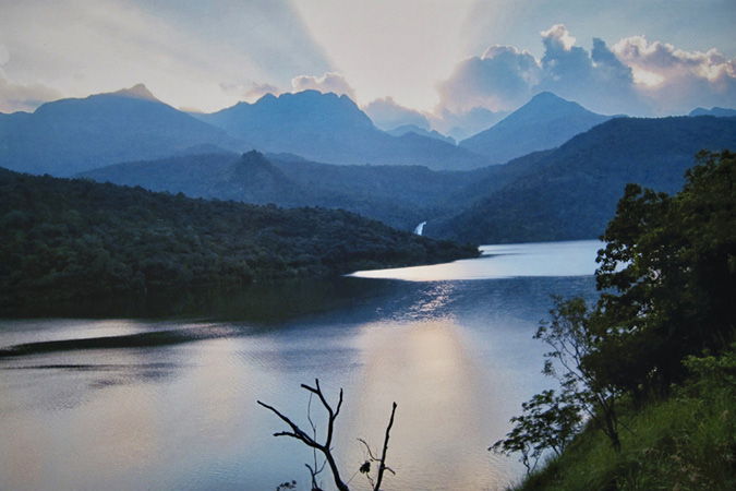 Looking out from the Papanasam upper dam in Tirunelveli in the state of Tamil Nadu, India is a view is of the Thambirabarani River at the foreground, sparkling in the light. Lush, green forests cover the rolling hill that rise from the shores of the river and extend to the horizon.