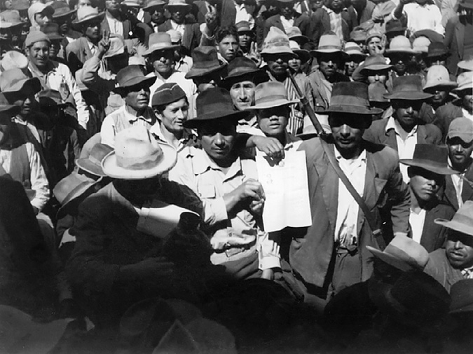 A man standing at the center of a crowd of peasants and next to an armed militiaman shows a document.