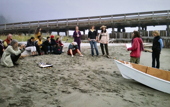 A group of people are seated on a sandy beach short with some standing at the far right of the semi-circle. Some of the observers have cameras or are simply looking forward. In front of the group on the beach is a white boat, the Lutra, waiting to be launched.