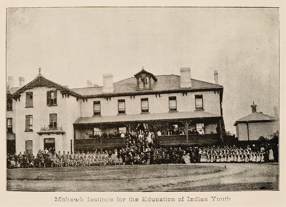 Black and white historical photograph. An imposing three story building with a long verandah dominates the scene. A large group of children are arranged in front of the building, in school uniforms, and separated into boys and girls, with adults on the verandah and stairs. The people are very small in front of the large building, so that their individual features cannot be distinguished. Some have musical instruments, including a large drum. Two Union Jacks hang from the verandah at the entrance to the school.
