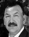 Black and white photograph: Portrait of a man with a moustache smiling and wearing a collared shirt and suit jacket. 