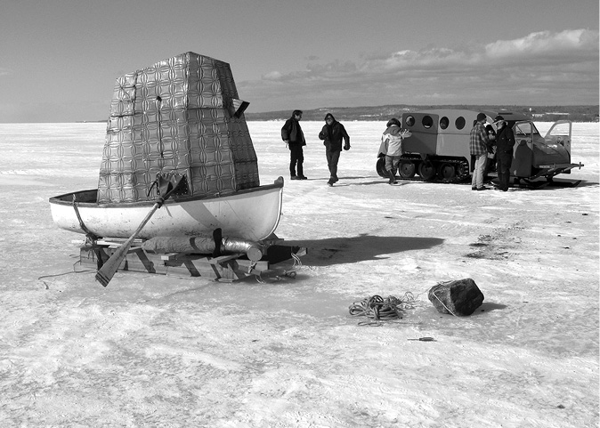 Black and white photograph, an irregular shaped camera obscura sits inside a canoe on a pallet jack on a frozen lake, with oars coming out of the camera obscura; a large snowmobile sits in the background; 5 people in winter clothing mill around the snowmobile, robe, a rock, and screwdriver sit in the foreground.