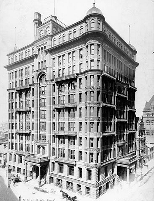 Black and white historical photograph. An imposing 12-storey early twentieth century skyscraper stands above the street, viewed from one corner. It is a square brick building in the Romanesque Revival style, with rounded corners and elaborate brownstone trim. There is a portico entrance on each of the two visible sides of the building. Small horses and carts can be seen at the foot of the building. A corner of Old Toronto City Hall is visible in the background.